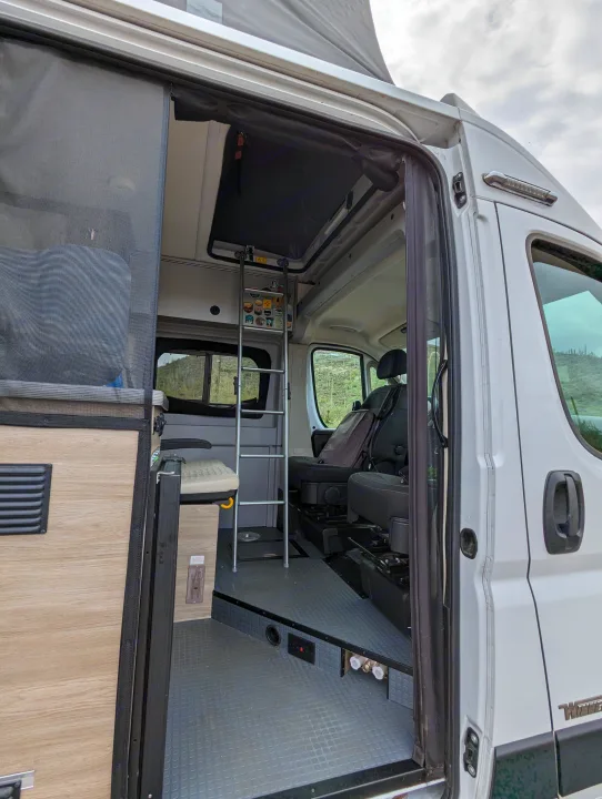 Open side door of an RV rental revealing a comfortable interior with bug screen, ladder to bed, and front seats.. Winnebago Solis 2021