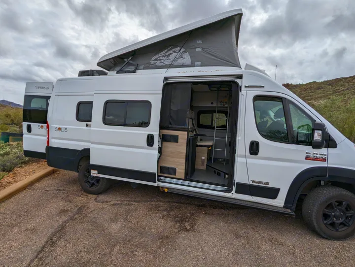 White Winnebago Solis camper van with pop-top raised, side door open, revealing interior. Perfect RV rental for desert adventures.. Winnebago Solis 2021