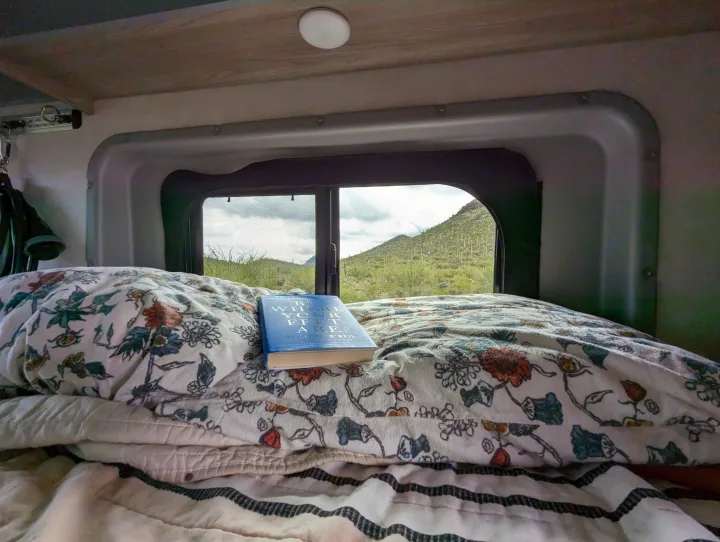 Cozy RV rental bed with a book, overlooking a scenic desert mountain landscape. Perfect for van life travel.. Winnebago Solis 2021