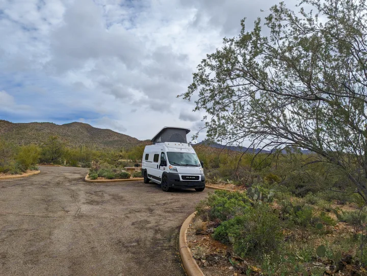 White pop-top RV rental parked at a desert campsite among saguaro cacti and mountains under cloudy sky.. Winnebago Solis 2021