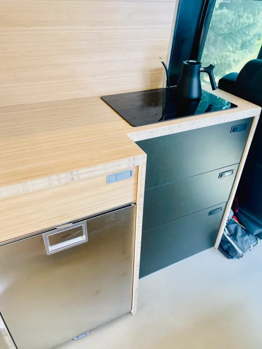 Modern RV rental kitchen with light wood counters, a Dometic fridge, built-in induction cooktop, and dark grey drawers.. Mercedes Sprinter 2500 2020
