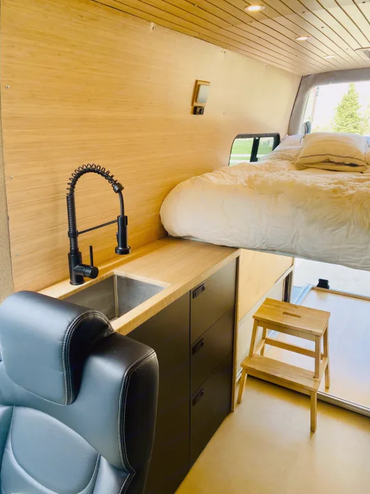 Cozy RV rental interior showing a raised bed, compact kitchen sink with black faucet, and wood paneling on walls and ceiling.. Mercedes Sprinter 2500 2020
