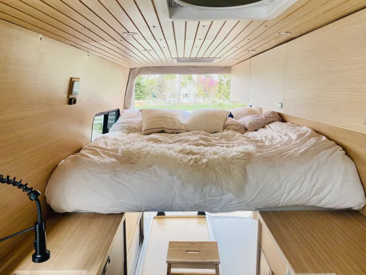 Interior of an RV rental camper van showing a large bed with white bedding, light wood walls, and a view outside.. Mercedes Sprinter 2500 2020