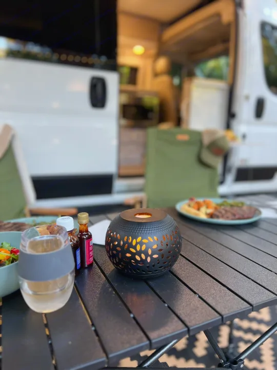 A cozy outdoor dinner by an RV rental: a black table holds a lit lantern, drink, and food, with green chairs and the RV's open door in the background.. Thor Sequence 2023