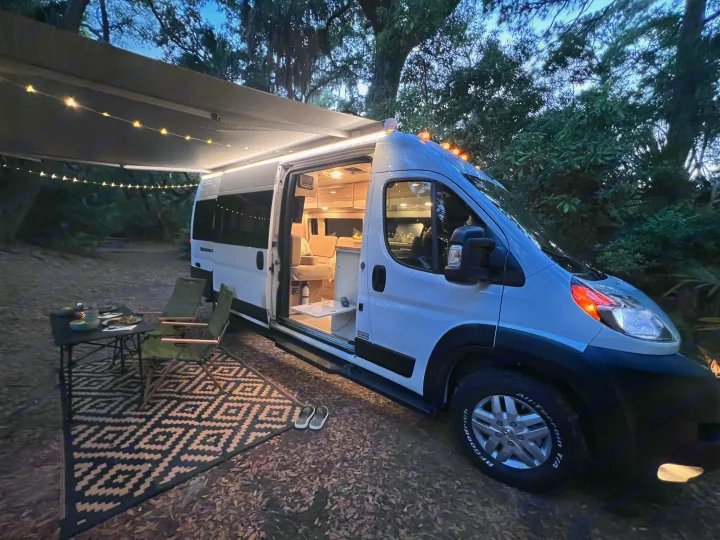This cozy camper van RV rental features an illuminated interior, string lights on the awning, and outdoor seating for evening relaxation.. Thor Sequence 2023