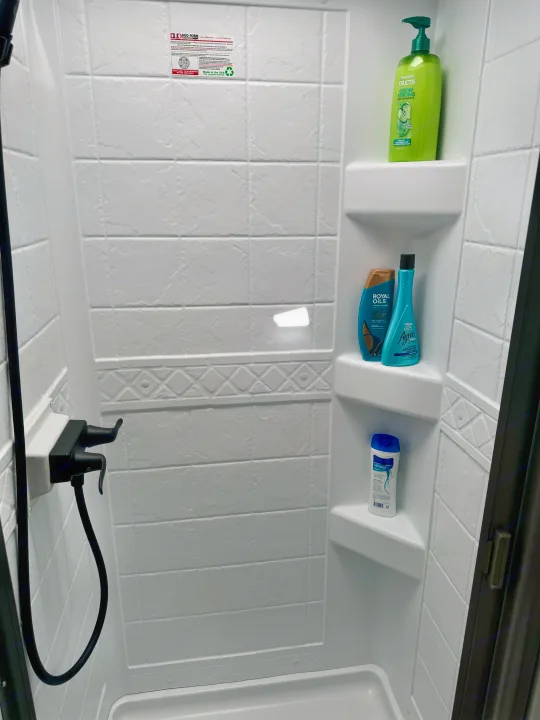 Compact shower in an rv rental, with white textured walls, shelves holding toiletries, and a modern black faucet.. Grand Design Imagine AIM 2024