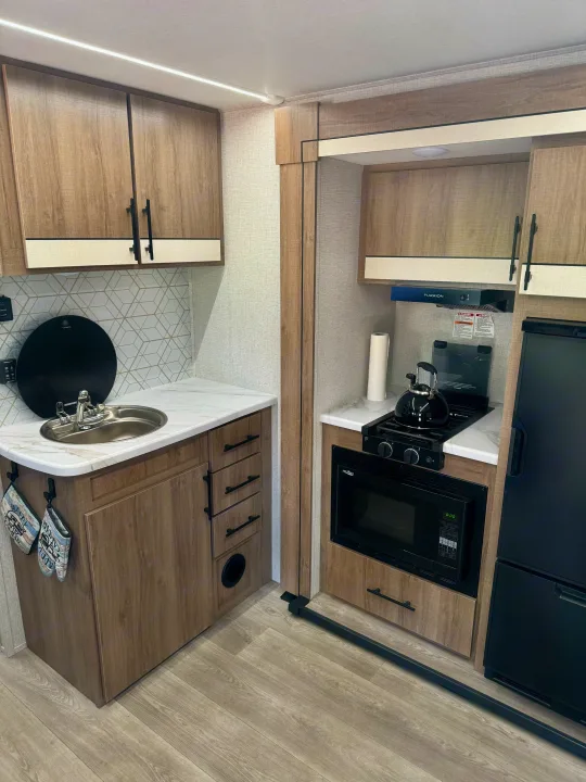 Well-equipped **rv rental** kitchen featuring a sink, stovetop, microwave, and refrigerator in light wood and white finishes.. Grand Design Imagine AIM 2024