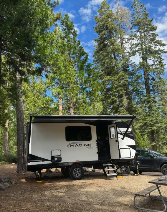 White Imagine Aim RV rental set up at a shaded forest campsite. Awning out, steps down for comfortable camping.. Grand Design Imagine AIM 2024