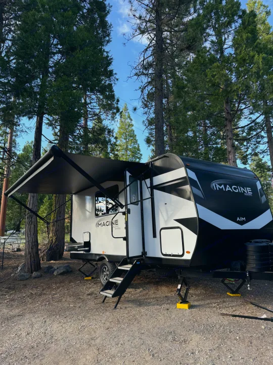 RV rental parked under tall trees with awning extended. This travel trailer is ready for a peaceful camping adventure.. Grand Design Imagine AIM 2024
