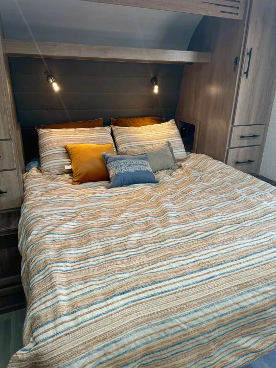 Cozy RV rental bedroom with a striped bedspread, pillows, built-in storage, and reading lights, ready for a comfortable stay.. Grand Design Imagine AIM 2024
