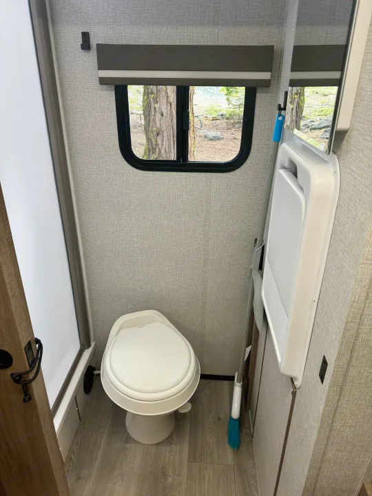 Experience comfort in this RV rental bathroom: compact toilet, window with trees, and wall-mounted fold-down table.. Grand Design Imagine AIM 2024