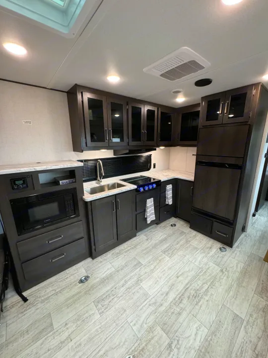 Modern RV rental kitchen featuring dark wood cabinets, light countertops, full appliances, and wood-look flooring.. Grand Design Grand Design Trailer 2024