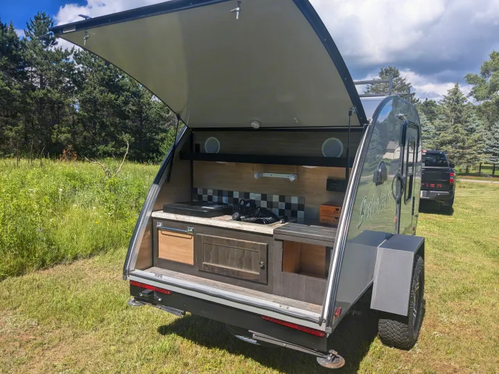 This rv rental, a grey teardrop camper, showcases its open rear hatch revealing a fully equipped outdoor kitchen setup.. Braxton Creek Bushwhacker 2021