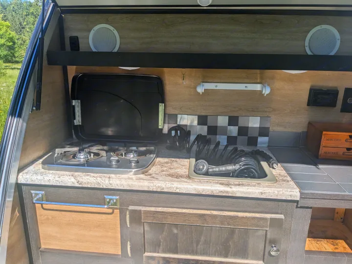 Compact RV rental outdoor kitchen: two-burner stove, sink with spray hose, speakers, and countertop storage.. Braxton Creek Bushwhacker 2021