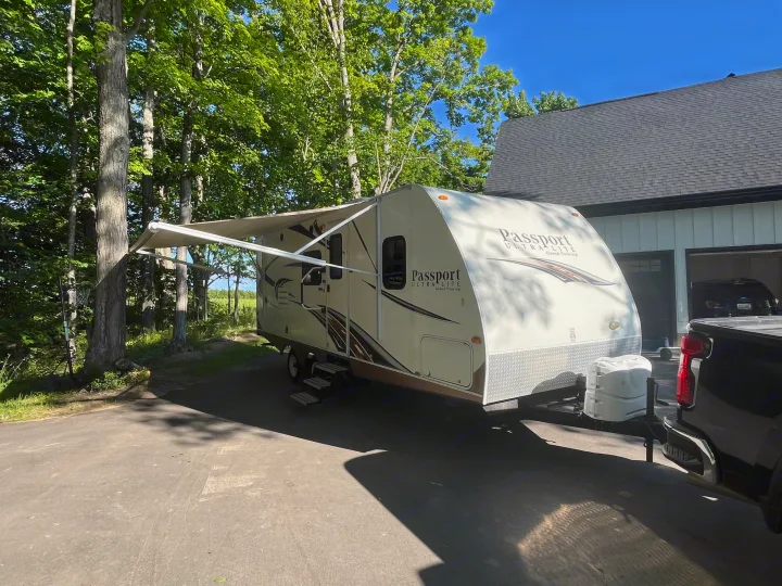 A white Passport Ultra-Lite travel trailer RV rental with its awning extended, parked on a sunny driveway next to a house.. Keystone RV Passport Ultra 2013
