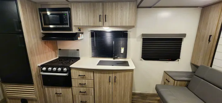 Modern RV rental kitchen featuring light wood cabinets, black appliances (microwave, stove, fridge), sink, and a compact dining bench.. Keystone RV Passport SL 2022
