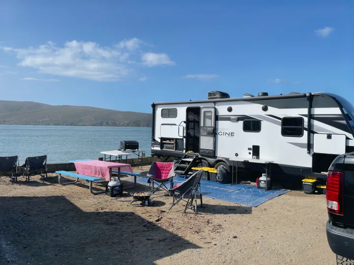 A modern rv rental parked waterside, set up for camping with a picnic table, grill, and chairs under blue skies.. Grand Design Imagine 2022