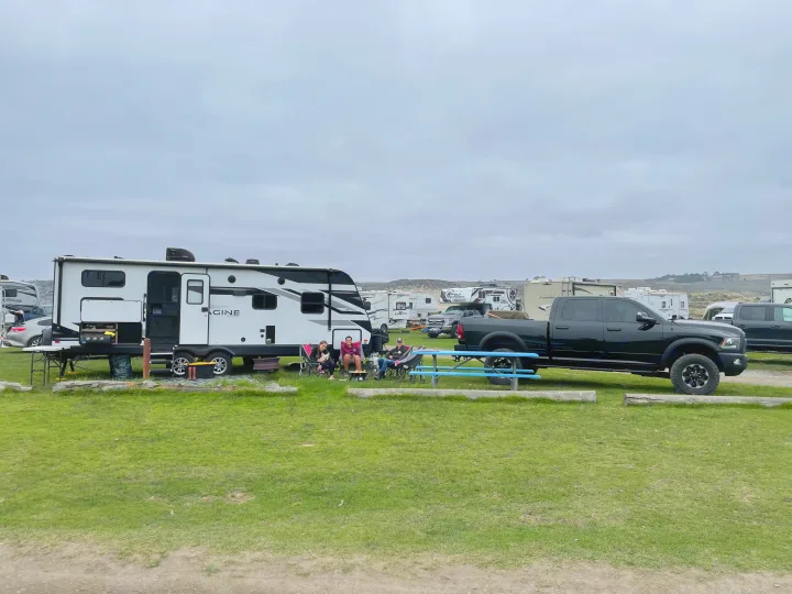 People and a dog relax outside a white RV rental & black truck at a grassy, overcast campground site.. Grand Design Imagine 2022