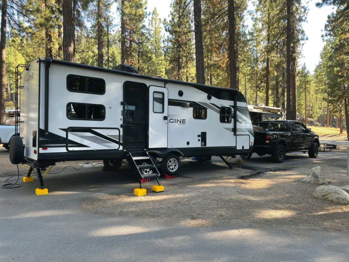 An Imagine Grand Design RV rental and a black pickup truck parked in a sunny forest campsite, with tall trees and dirt ground.. Grand Design Imagine 2022