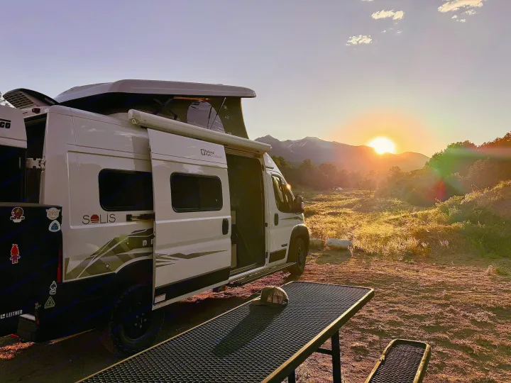 A white RV rental camper van with its pop-top extended at sunrise, parked with a stunning mountain view.. Winnebago Solis 2023
