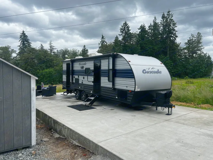 Parked Cascade travel trailer RV rental on a concrete pad with outdoor furniture, surrounded by nature under an overcast sky.. Forest River Cherokee Grey Wolf 2021