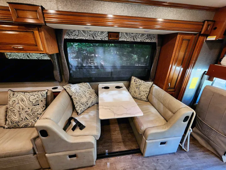 Interior of an RV rental: a bright dinette with patterned pillows, beige benches, table, wood cabinets, and window views.. Fleetwood Bounder 2018