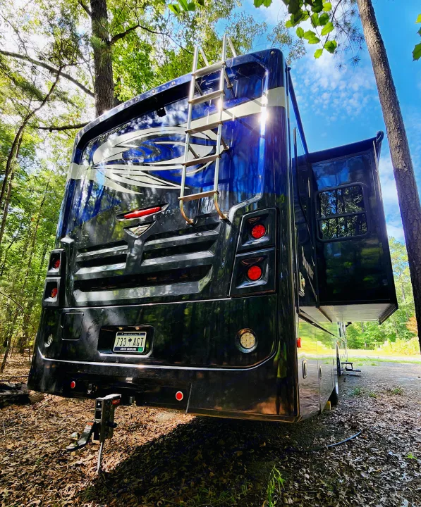 A sleek blue-black RV rental, with an extended slide-out and rear ladder, is perfectly parked in a sunny, serene wooded campsite.. Fleetwood Bounder 2018