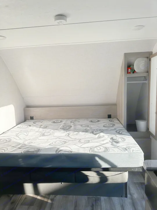 RV rental bedroom: A bed with a grey swirly mattress, light wood headboard, and storage cabinet with games. Sunlight visible.. Dutchmen Coleman Light 2022