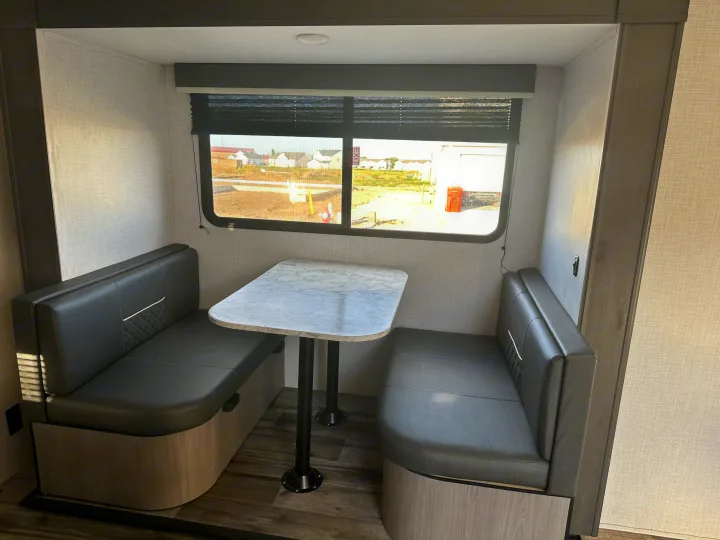 Inside an RV rental, a cozy dinette booth features dark upholstered seating, a white marble-pattern table, and a large window.. Dutchmen Coleman Light 2022