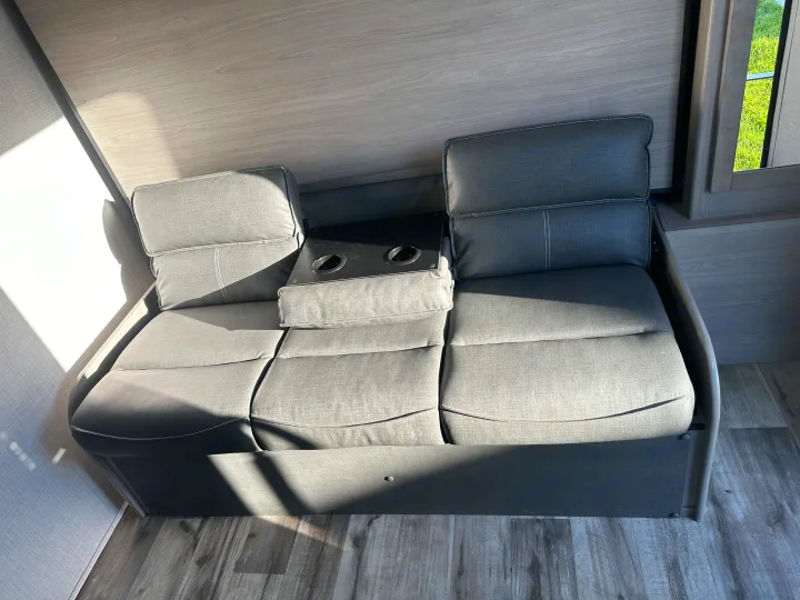 Gray fabric RV rental sofa with integrated cup holders. Sunlight brightens the modern interior, showing wood-grain walls.. Dutchmen Coleman Light 2022