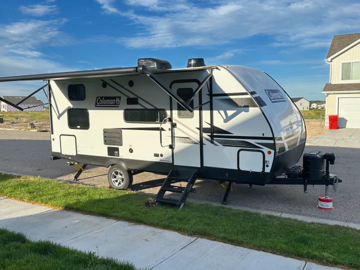 A Coleman travel trailer, potentially an rv rental, is parked with its awning extended in a residential area under a blue sky.. Dutchmen Coleman Light 2022