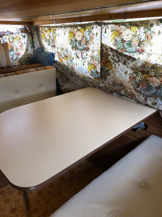 Inside an RV rental: a bright dinette with tufted white benches, a long table, and vintage floral curtains.. Vintage Trailers Skylark 180 1979