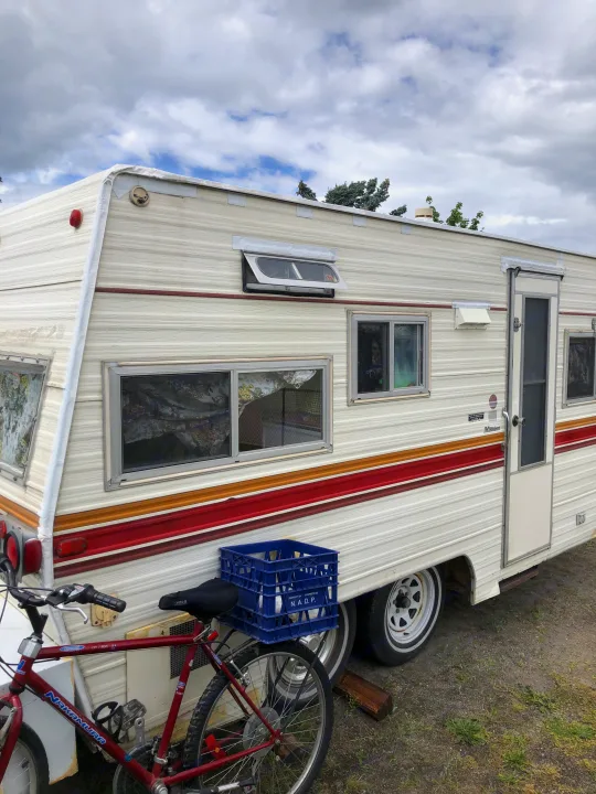 Cream-colored camper RV rental with retro stripes, parked next to a red bicycle, under a cloudy sky.. Vintage Trailers Skylark 180 1979