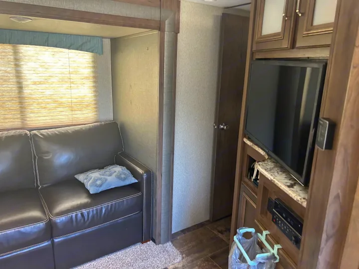 Inside an RV rental: a cozy living area with a dark leather couch, bright window, and built-in entertainment system with TV.. Keystone RV Passport Grand Touring 2018