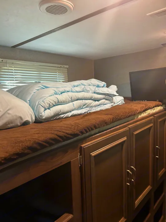 RV rental interior: A comfy bunk bed with light blue comforter, brown blanket, integrated cabinets, and TV.. Keystone RV Passport Grand Touring 2018
