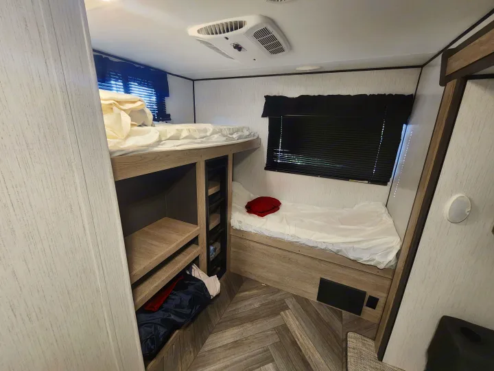 Inside an RV rental: a compact bunk bed sleeping area with storage shelves, ceiling AC, and wood-look flooring.. Heartland RVs Prowler 2022