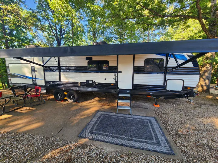 A white & black RV rental is parked at a sunny campsite with an extended awning, steps, outdoor rug, picnic table, and chairs.. Heartland RVs Prowler 2022