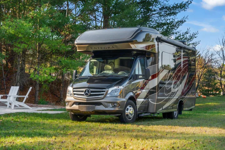 Ready for adventure, this dark brown Mercedes-Benz RV rental is parked on a green lawn with a lush treeline.. Entegra Coach Qwest 2018