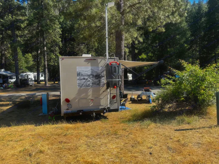 RV rental parked at a sunny campsite with an awning extended, surrounded by tall pine trees.. Coachmen Leprechaun 270QB 2019