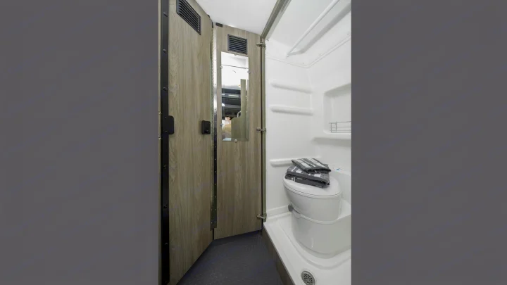 This RV rental bathroom features a compact white toilet, shower, and a wooden door with a mirror, designed for practical travel.. Winnebago Solis 2021