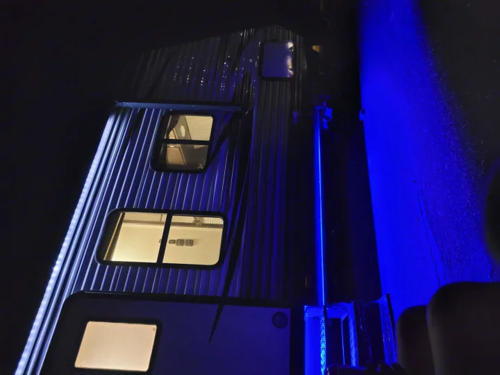 Side of an **rv rental** at night with bright interior lights from windows & blue exterior illumination. Corrugated siding.. Heartland RVs Pioneer 2020