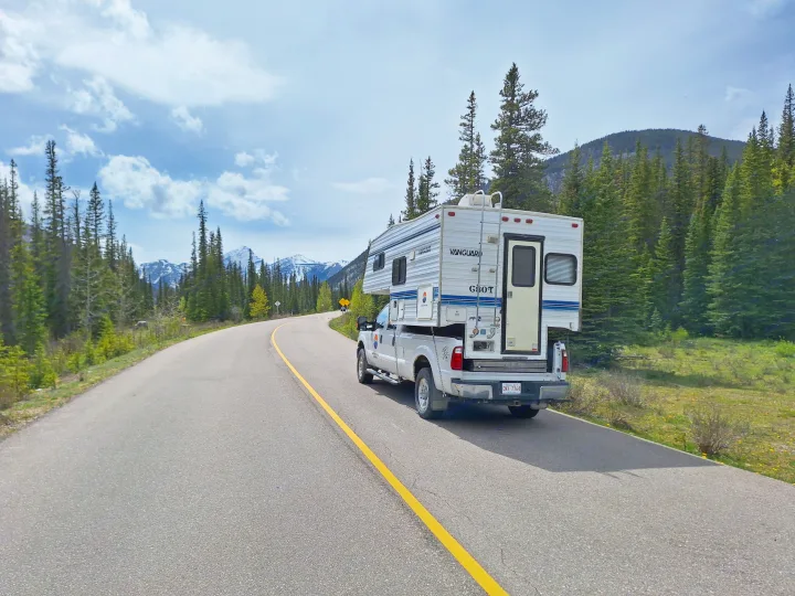 RV rental truck camper parked on a winding mountain road, surrounded by pine forests and snow-capped peaks, ready for adventure.. Glacier Campers Truck Camper 2016