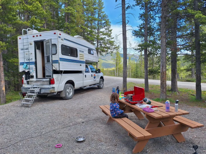 RV rental adventure: a child sits at a picnic table with a camp stove, next to a truck camper, surrounded by trees and mountains.. Glacier Campers Truck Camper 2016