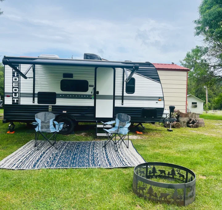 An RV rental set up on a grassy campsite with an outdoor rug, two chairs, and a decorative fire pit.. Keystone RV Hideout 2022