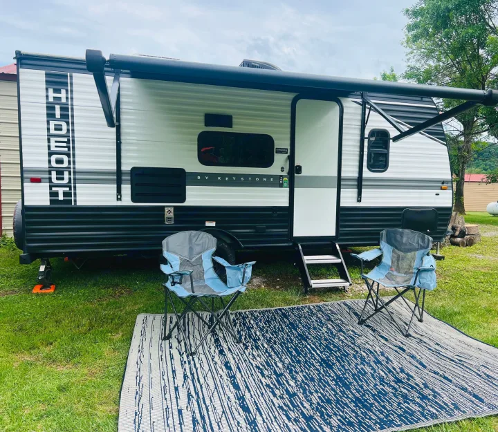Comfortable rv rental setup: a travel trailer with extended awning, two camping chairs, and an outdoor rug on the grass.. Keystone RV Hideout 2022