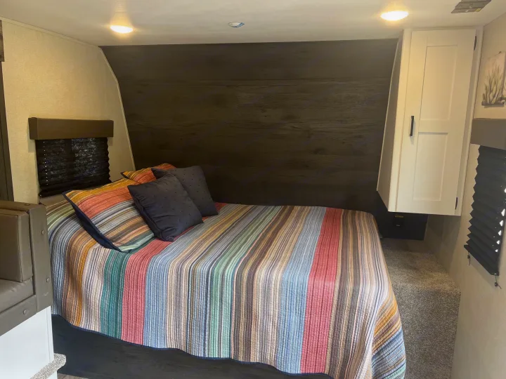 Spacious RV rental bedroom with a large bed, vibrant striped quilt, dark wood wall, and white cabinet, offering comfort.. Keystone RV Hideout 2022