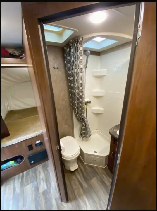 RV rental interior showing a compact bathroom with shower, toilet, and sink. A sleeping bunk is partially visible.. Jayco Jay Feather 2017