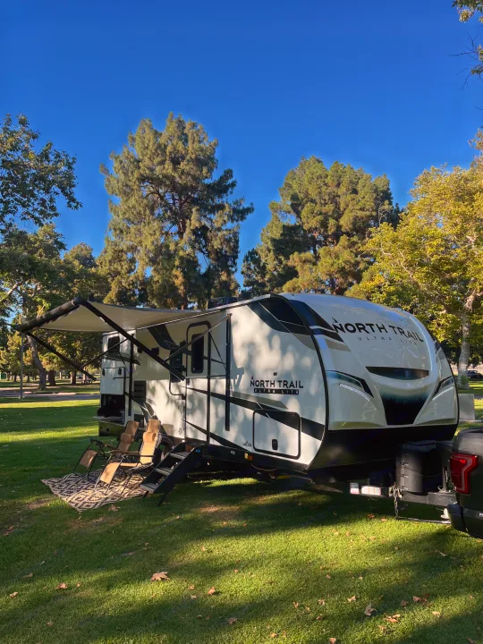 White and black RV rental, a North Trail Ultra Lite, parked on lush grass with its awning extended and chairs set up in a sunny park.. Heartland RVs North Trail 2022