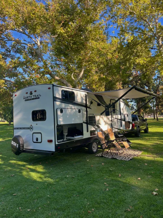 RV rental fun! This white travel trailer is parked on green grass with an extended awning, outdoor kitchen, and chairs.. Heartland RVs North Trail 2022