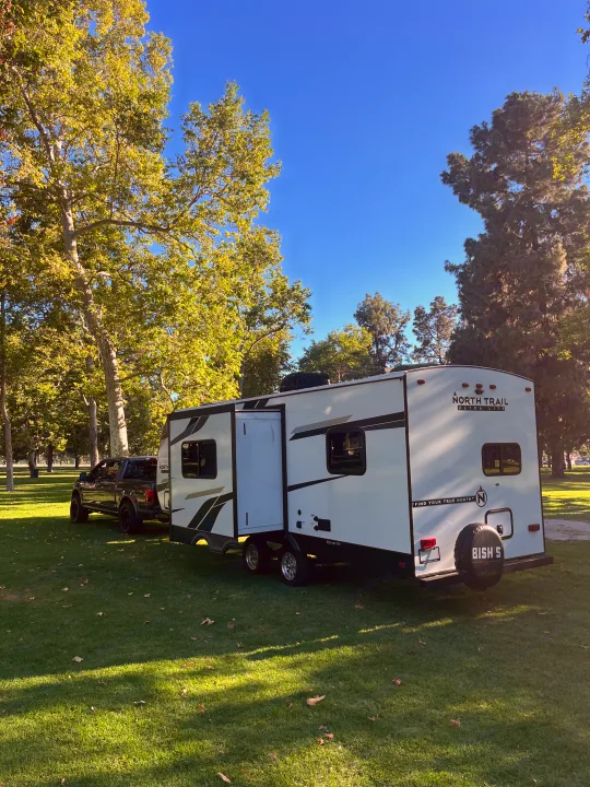 Modern travel trailer RV and truck parked on green grass with trees under a blue sky. Ideal for a scenic outdoor rv rental getaway!. Heartland RVs North Trail 2022